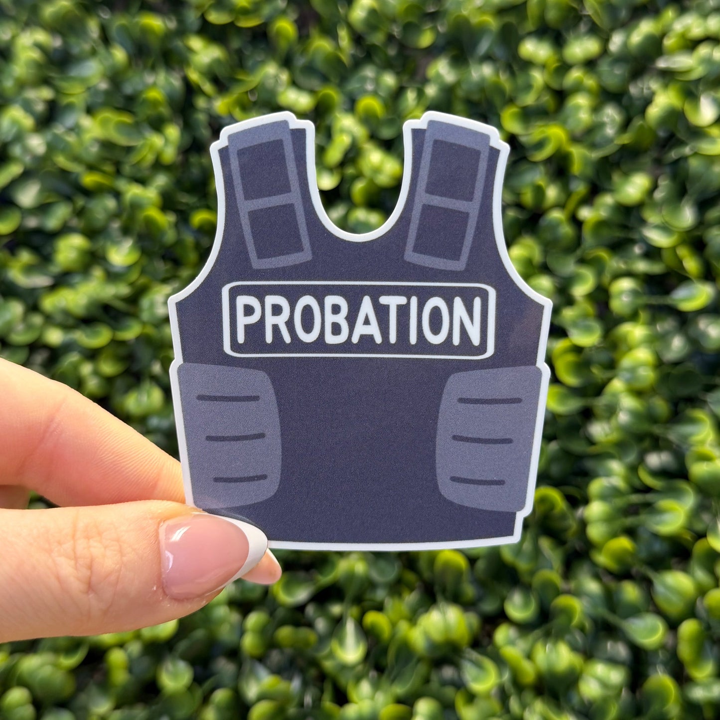 Sticker of a bulletproof vest labeled 'Probation' held in front of green foliage.