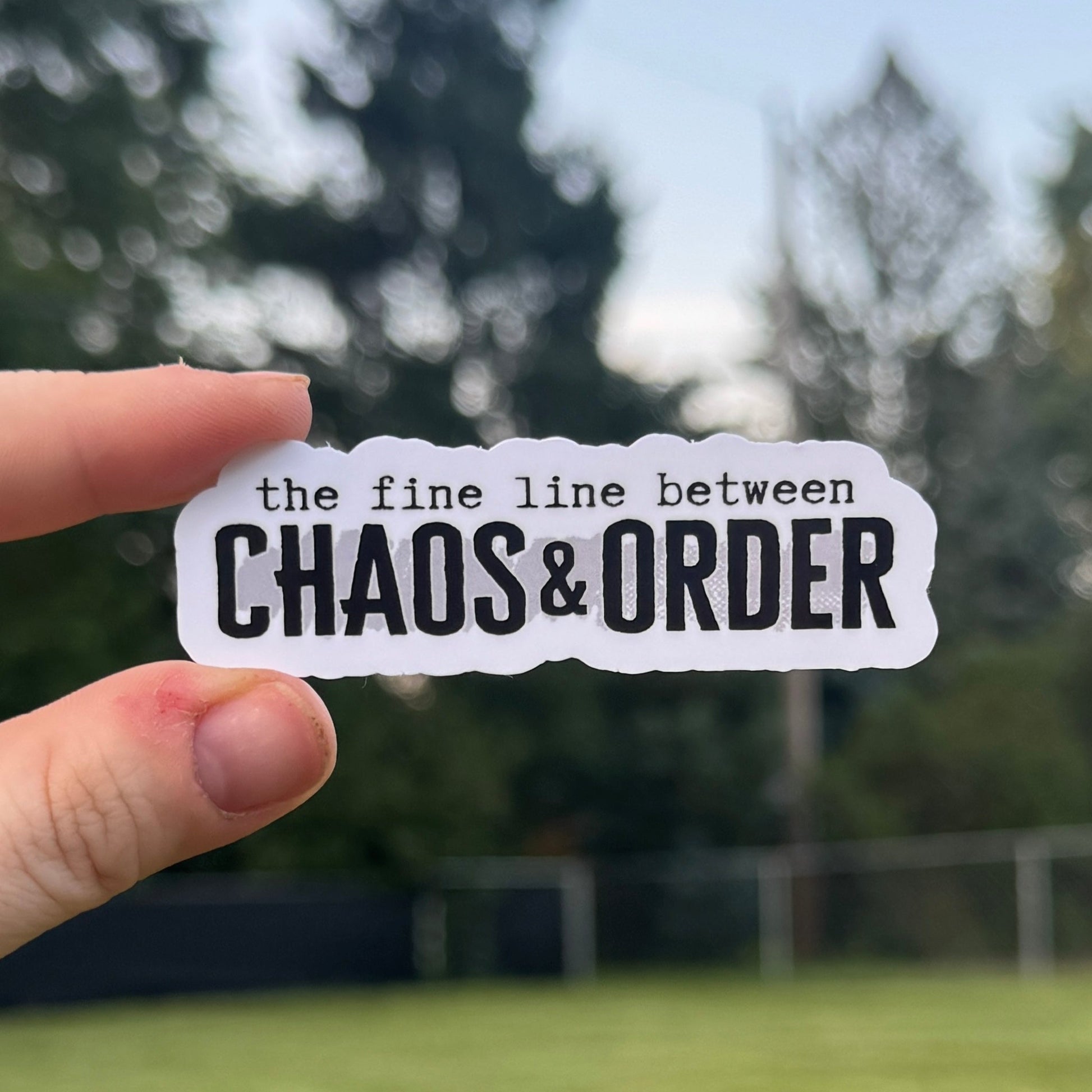 Hand holding a sticker with 'the fine line between CHAOS & ORDER' text against a blurred outdoor background.