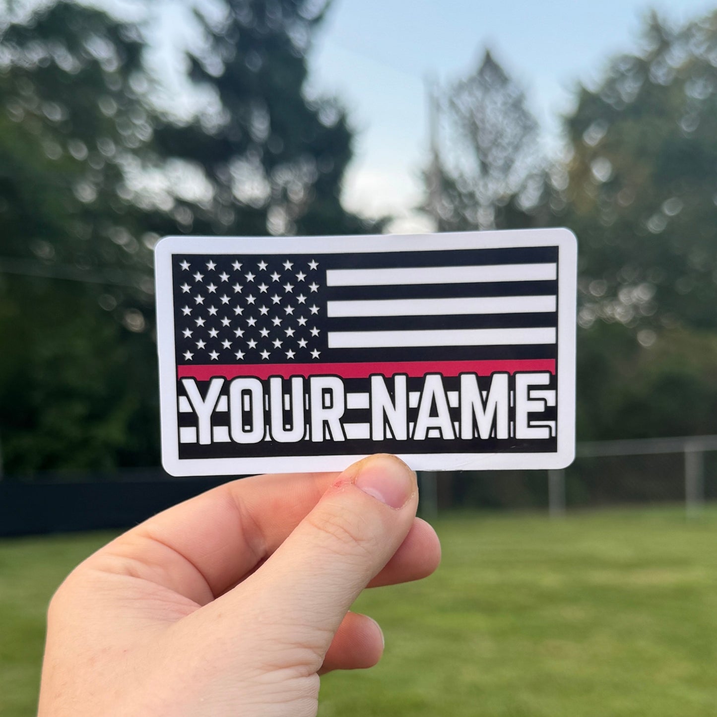Hand holding a customizable American flag sticker with 'YOUR NAME' text outdoors.
