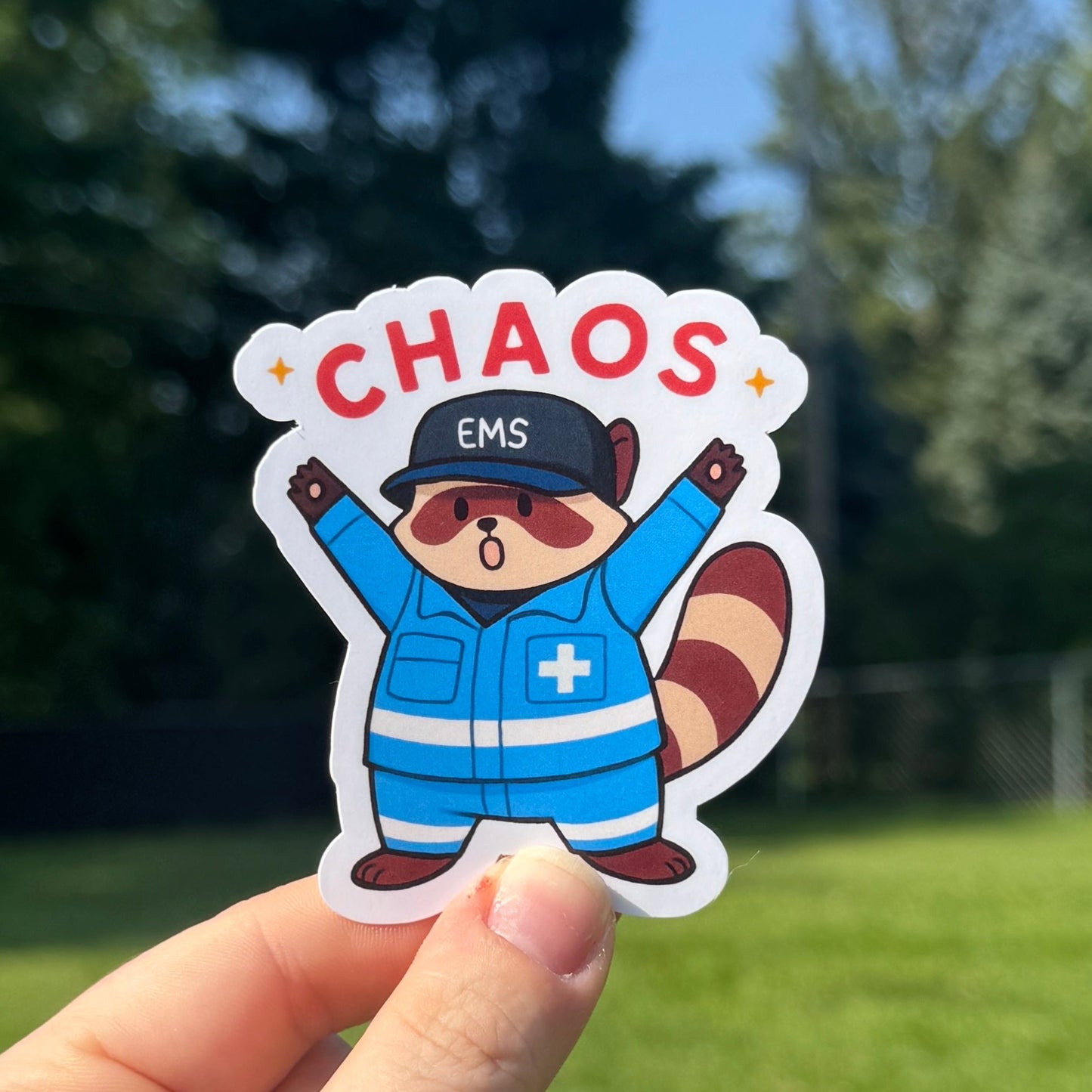 Hand holding a sticker with a cartoon character labeled 'Chaos EMS' against a green outdoor background.