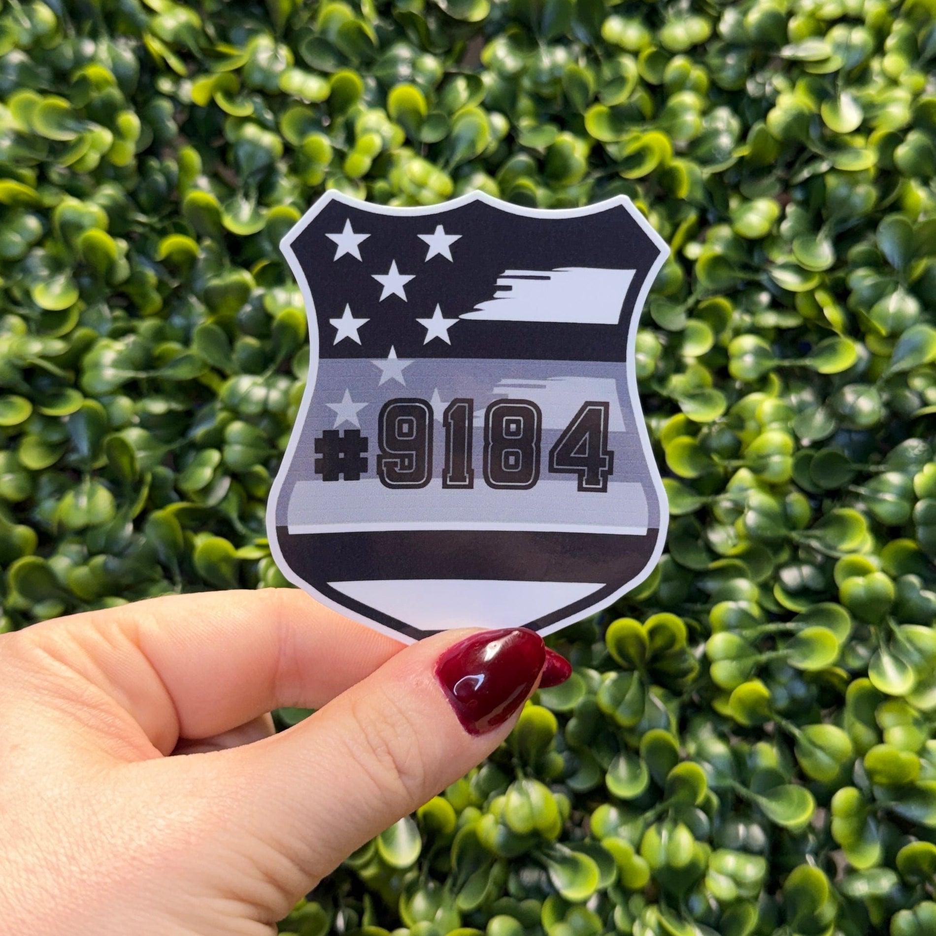 Hand holding a shield-shaped sticker with stars and numbers against a green leafy background