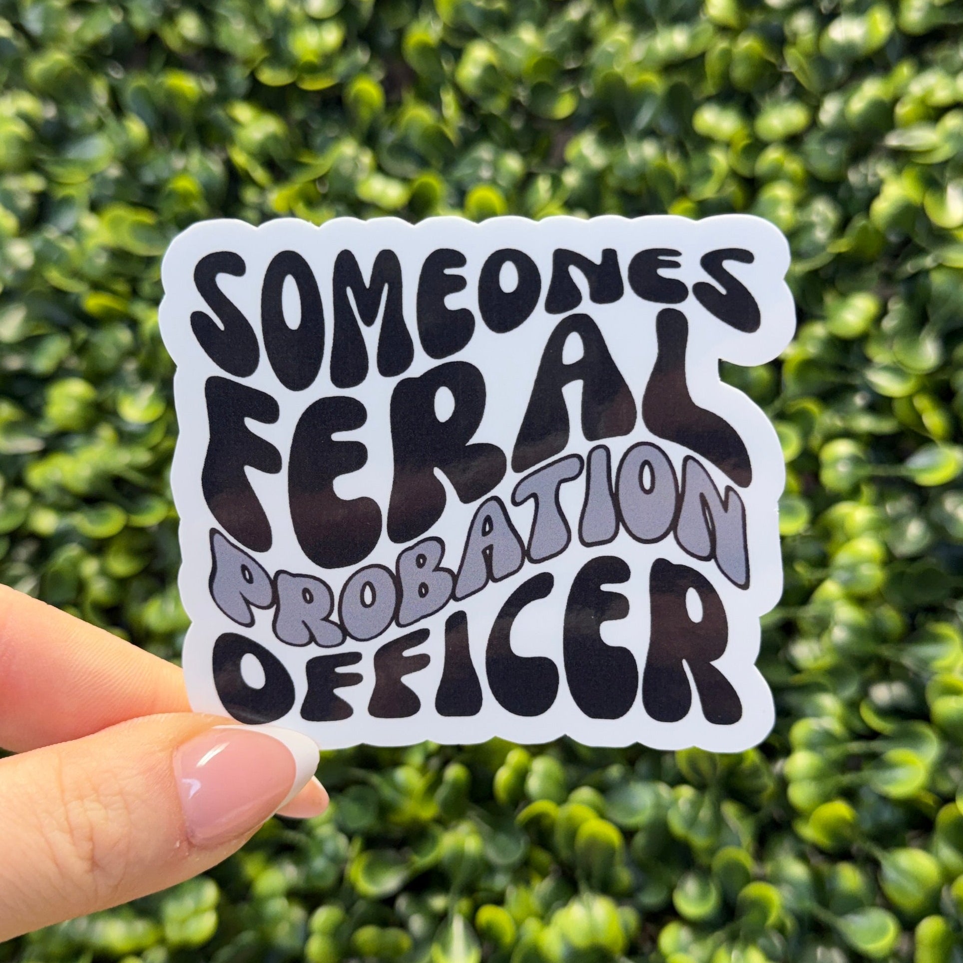 Hand holding a sticker with text against a green hedge background