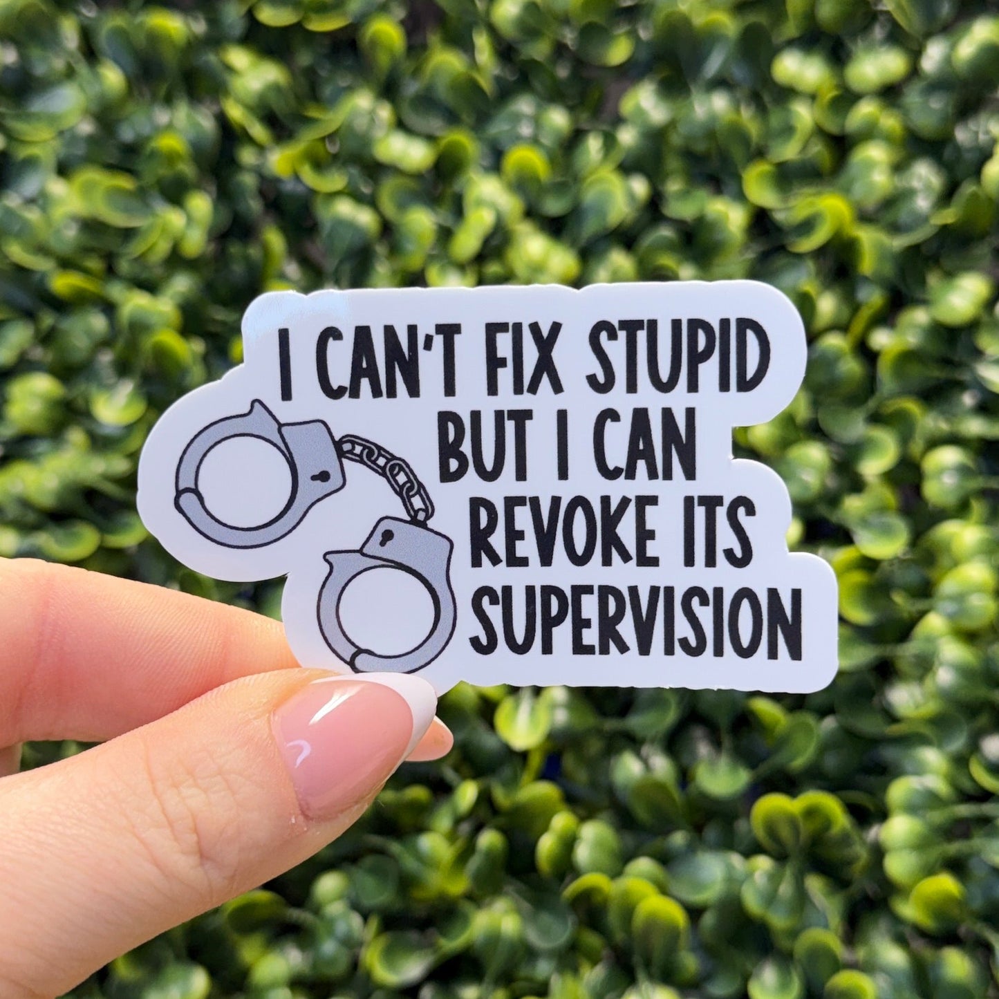 Hand holding a sticker with text and handcuffs design against a green bush background