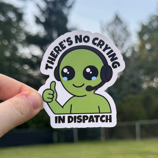 Hand holding a sticker with a cartoon character and text 'There's no crying in dispatch' against a blurred outdoor background.