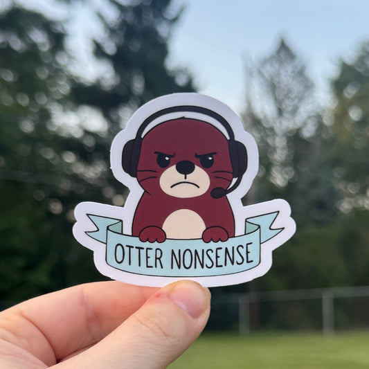 Hand holding a sticker with an otter wearing headphones and 'Otter Nonsense' text, outdoors.