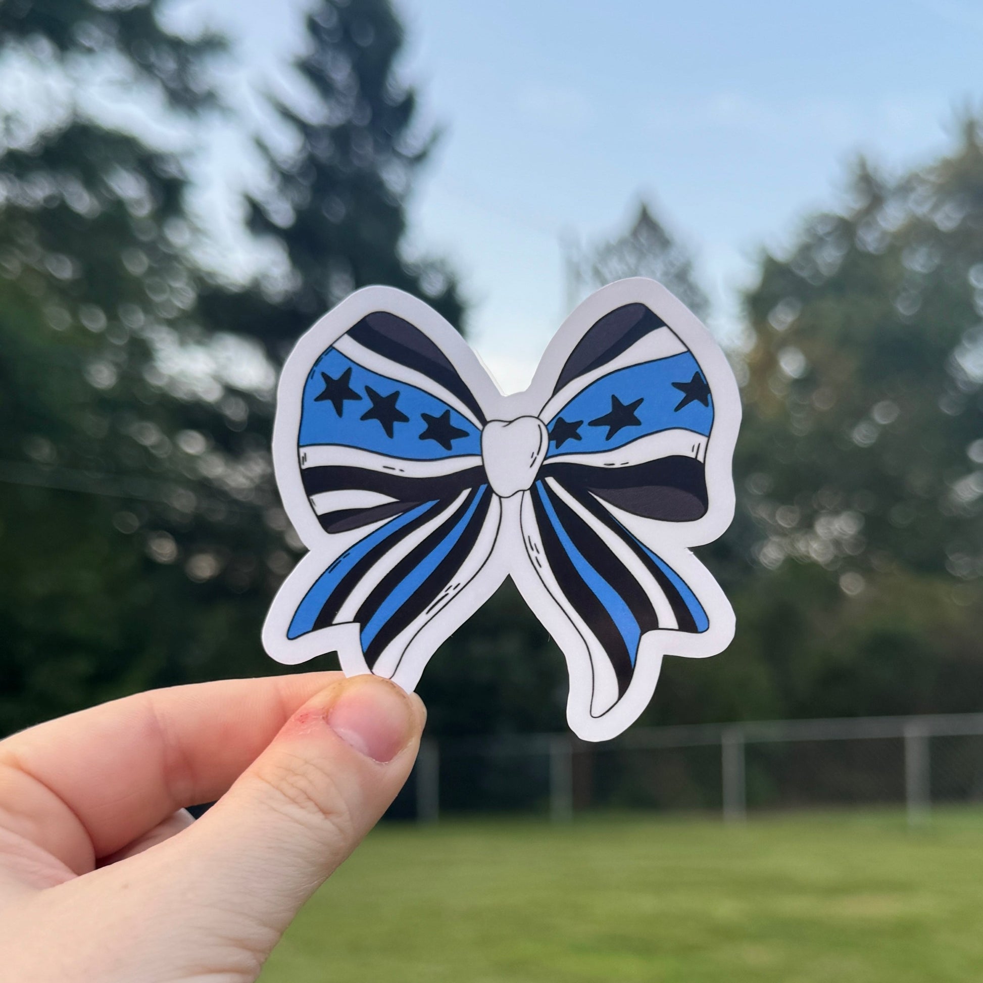 Hand holding a blue and white striped bow sticker outdoors with trees in the background